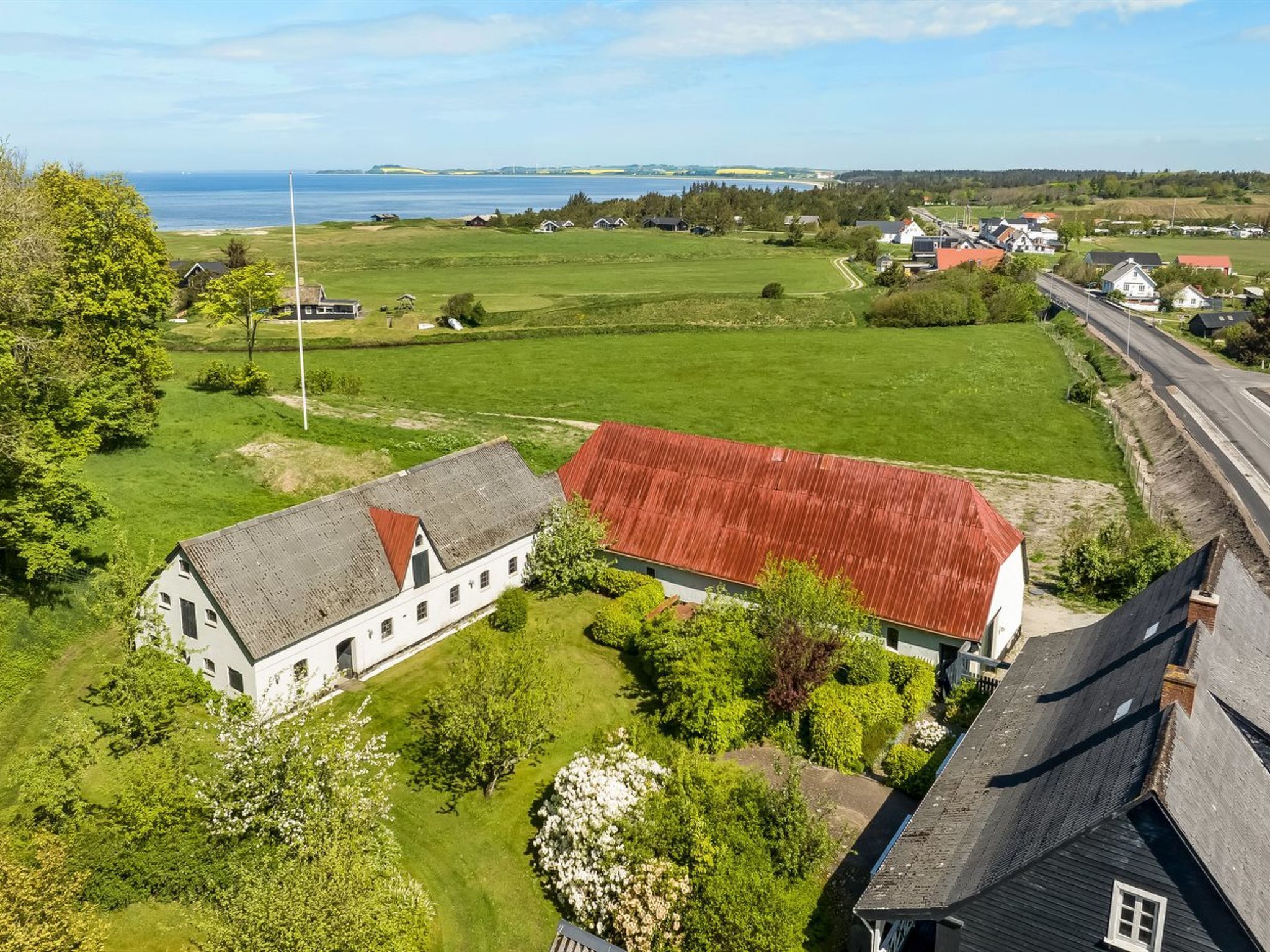 A rural landscape with a white building with a red roof, surrounded by green fields and a body of water in the background.