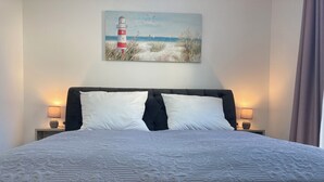 A neatly made bed with a patterned bedspread, two white pillows, and a headboard. A painting of a lighthouse and beach scene hangs above the bed. - "bijoux - Juwel" im Haus Meeresjuwel! (Dahme)