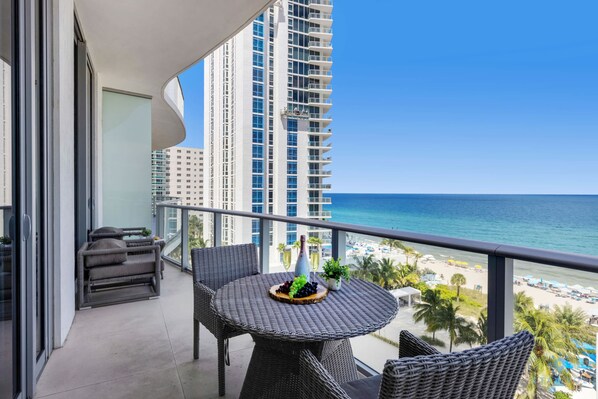 Un balcon avec une table ronde, des chaises en osier et une vue sur l’océan et la plage.