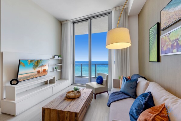 A modern living room with a television, a wooden coffee table, a sofa with colorful cushions, and a large window offering a view of the sea.