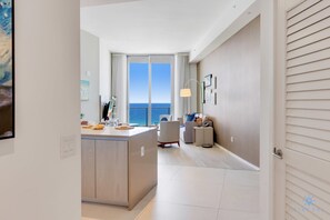 A modern hotel room with a large window offering a view of the sea, a kitchenette, a seating area with a sofa and armchairs, and a small side table. - Rooftop Pool –Oceanview Balcony| Beachfront Resort (Hollywood)