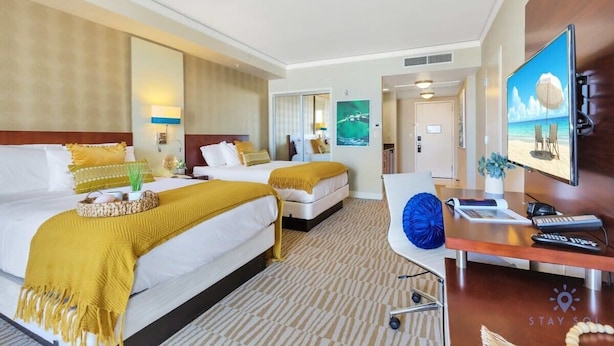 Ein Hotelzimmer mit zwei Betten, einem Schreibtisch mit Computer, einem Fernseher und Blick auf den Strand.