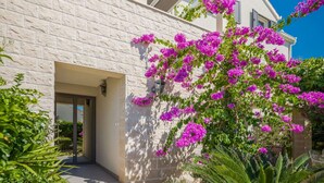 A modern building with a stone facade, a covered entrance, and vibrant bougainvillea flowers. - Apartment Oaza (Supetar)