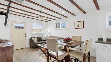 Un comedor con una mesa de madera, cuatro sillas y un ventilador de techo. Hay una zona de cocina con armarios blancos y un ventanal.