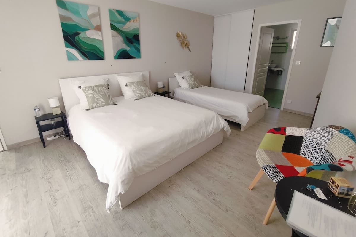 Ein Schlafzimmer mit zwei Betten, einem kleinen Tisch und einem Stuhl. An der Wand hängen abstrakte Gemälde, ein Nachttisch und eine Tür zu einem anderen Raum.