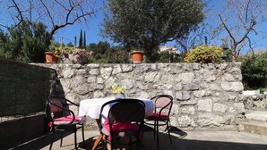 An outdoor patio with a stone wall, two tables with white tablecloths, and black metal chairs with red cushions.