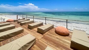 A wooden deck with beige cushioned seating and a railing overlooking a beach. - LaPili Residence (Kiwengwa)
