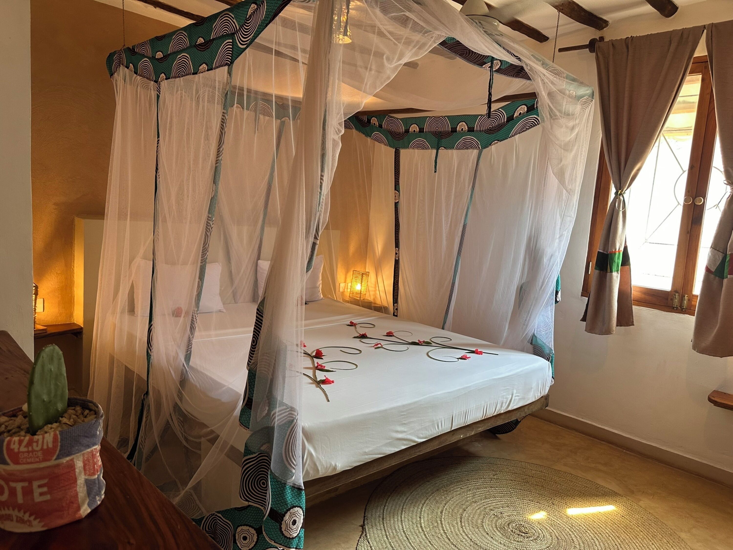 Una cama con dosel y cuatro columnas, con cubrecama blanco y flores rojas, un tapete tejido y un cactus en maceta al lado.