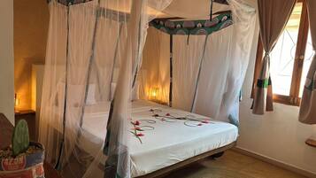 Una cama con dosel y cuatro columnas, con cubrecama blanco y flores rojas, un tapete tejido y un cactus en maceta al lado.