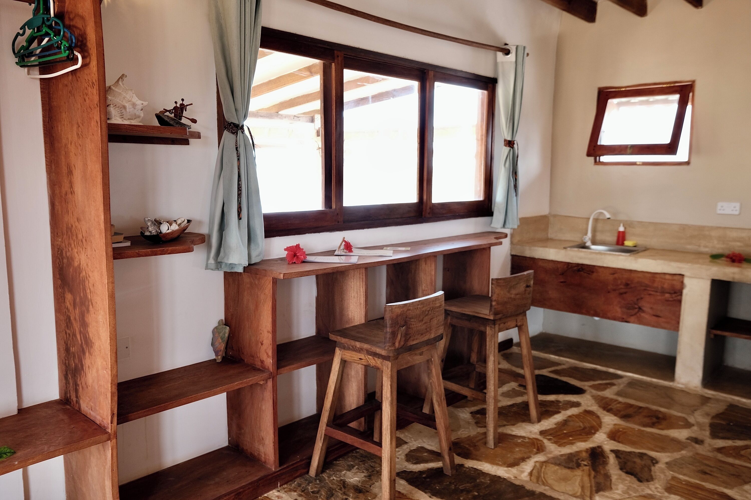 A rustic kitchen with wooden shelves, a stone floor, and a window with curtains.