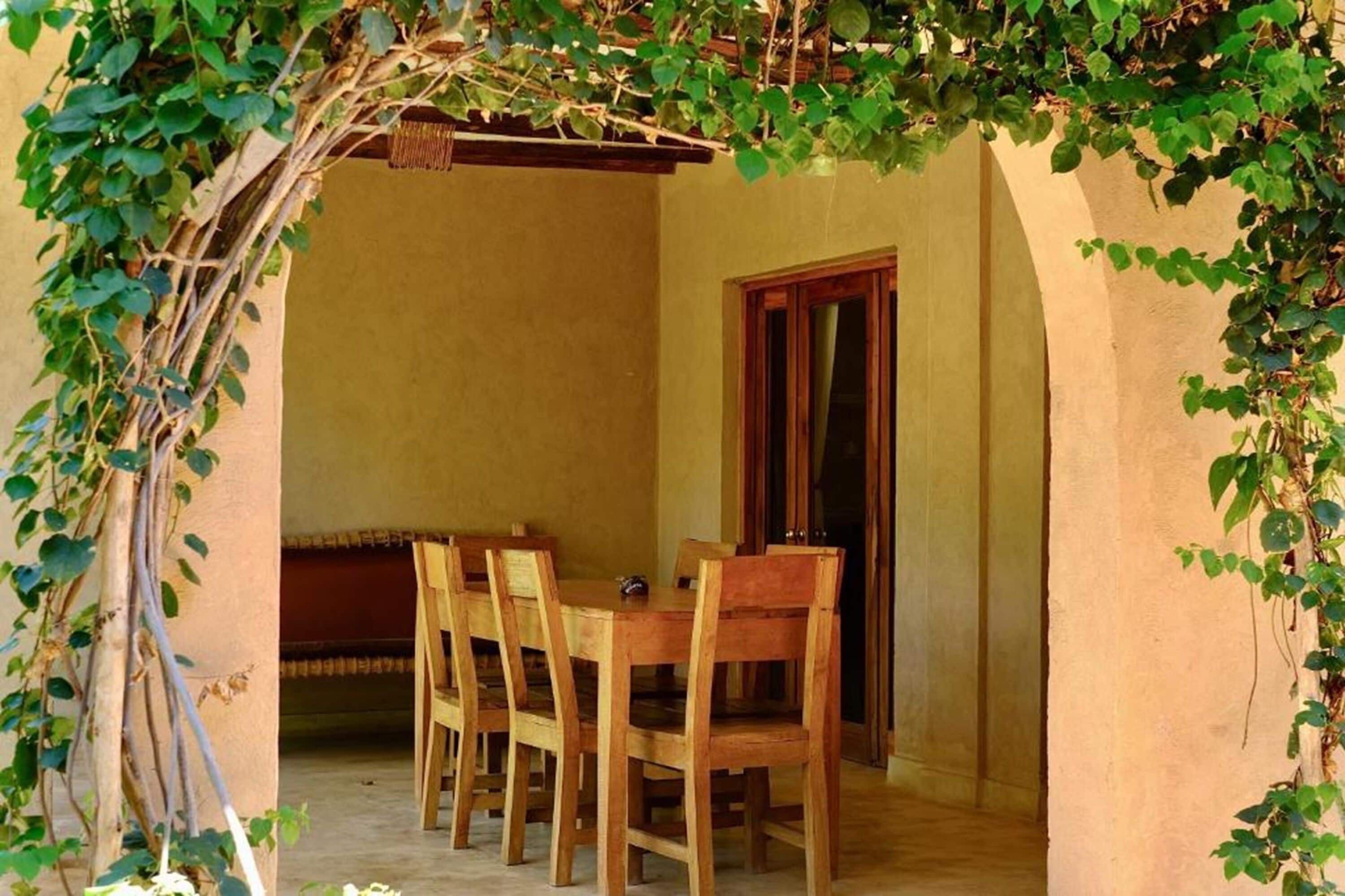 Uma área de refeições com mesa e cadeiras de madeira, uma porta de madeira e uma parede com plantas trepadeiras.