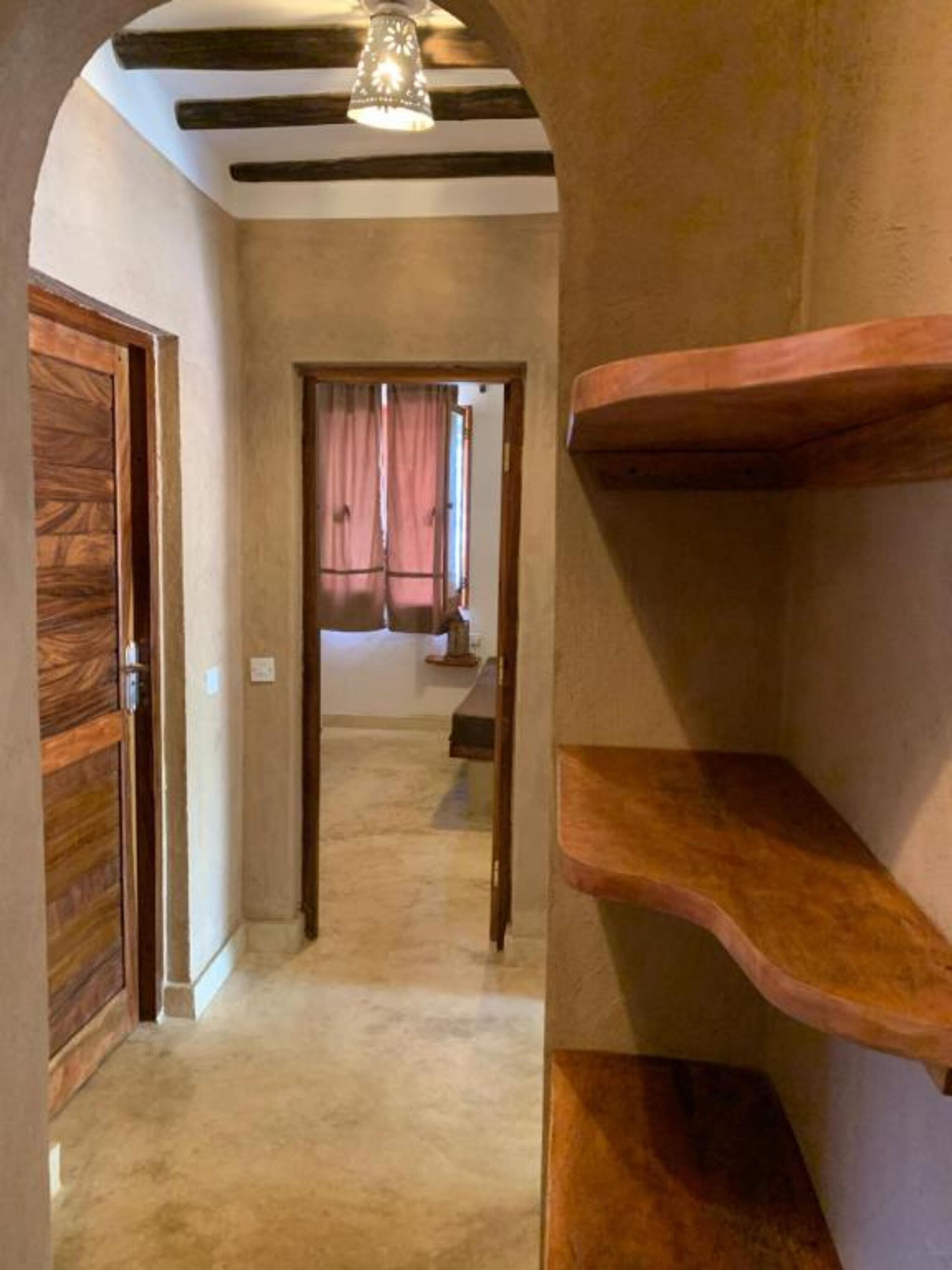 A hallway with wooden doors and shelves, leading to a room with a window and curtains.