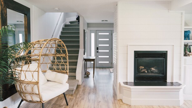 A modern living room with a rattan chair, a fireplace, and a staircase leading to another room. - Sandbanks Escape (5 min drive) on Sheba’s Island | Pet-Friendly Cottage (Prince Edward)