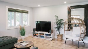 A modern living room with a flat-screen TV, a green sofa, a wicker chair, and a bookshelf. - Sandbanks Escape (5 min drive) on Sheba’s Island | Pet-Friendly Cottage (Prince Edward)