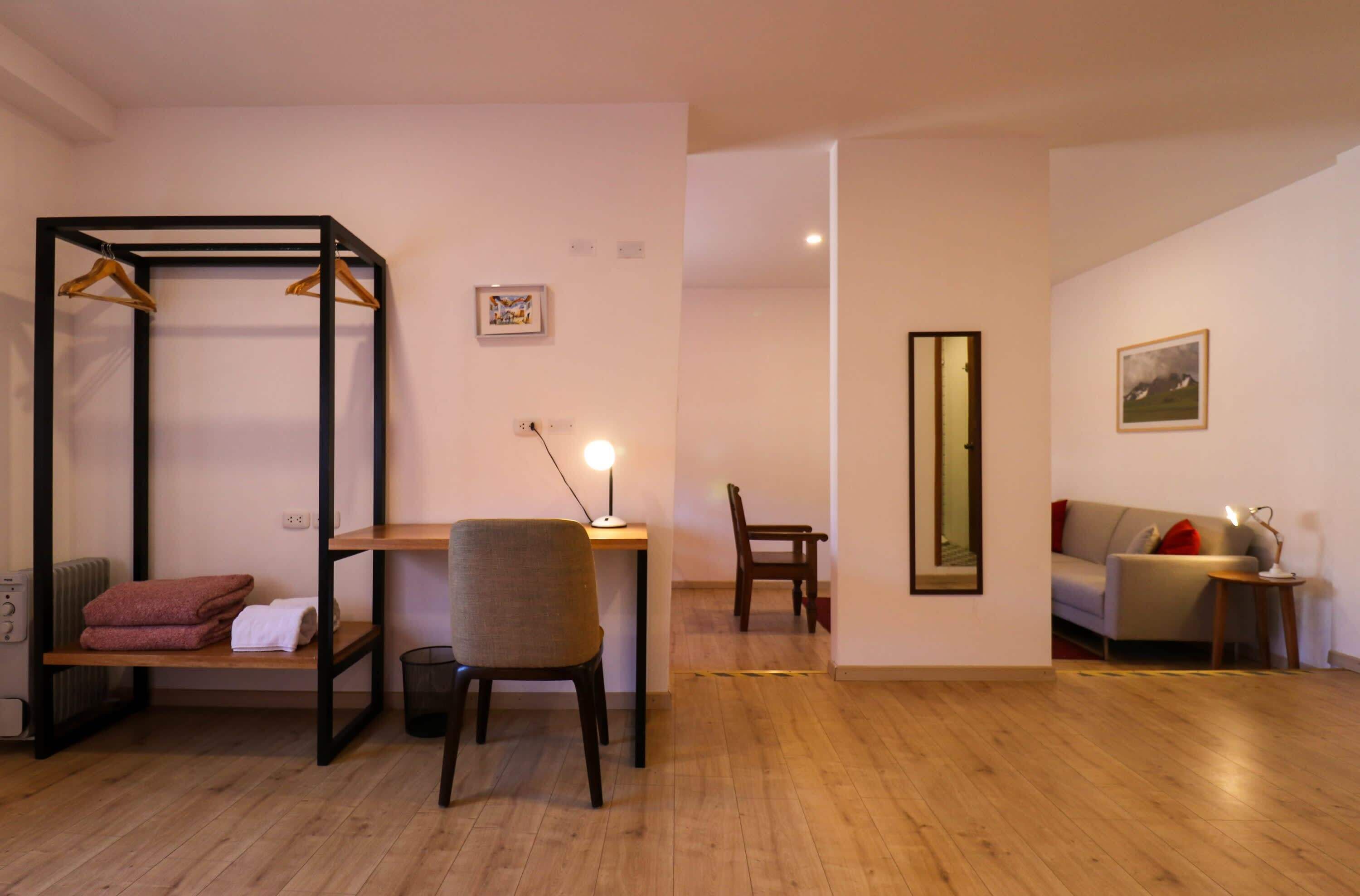 A room with a wooden floor, a black metal wardrobe with hangers, a chair, a desk with a lamp, a sofa, and a mirror on the wall.