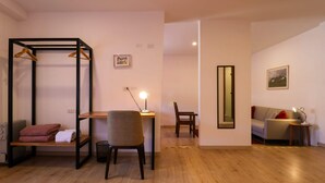 A room with a wooden floor, a black metal wardrobe with hangers, a chair, a desk with a lamp, a sofa, and a mirror on the wall.