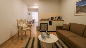 Ein gemütlicher, kleiner Wohnbereich mit einem Holzstuhl, einem braunen Sofa, einem kleinen Couchtisch und einer Küchenzeile, inklusive Kaffeemaschine und Kühlschrank.