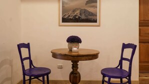 Een kamer met een ronde houten tafel, twee paarse stoelen, een ingelijste afbeelding van een berg en een vaas met paarse bloemen.