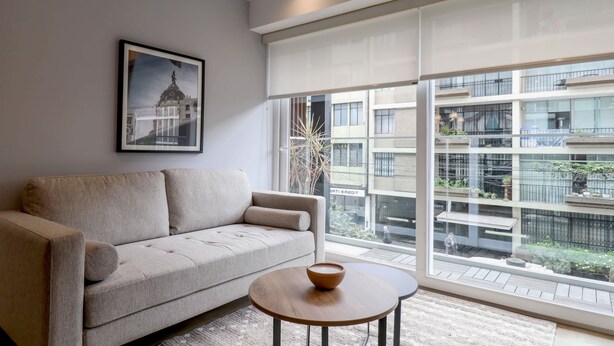 Una sala de estar moderna con un sofá, una mesita redonda y un amplio ventanal con vistas a un edificio y zonas verdes.