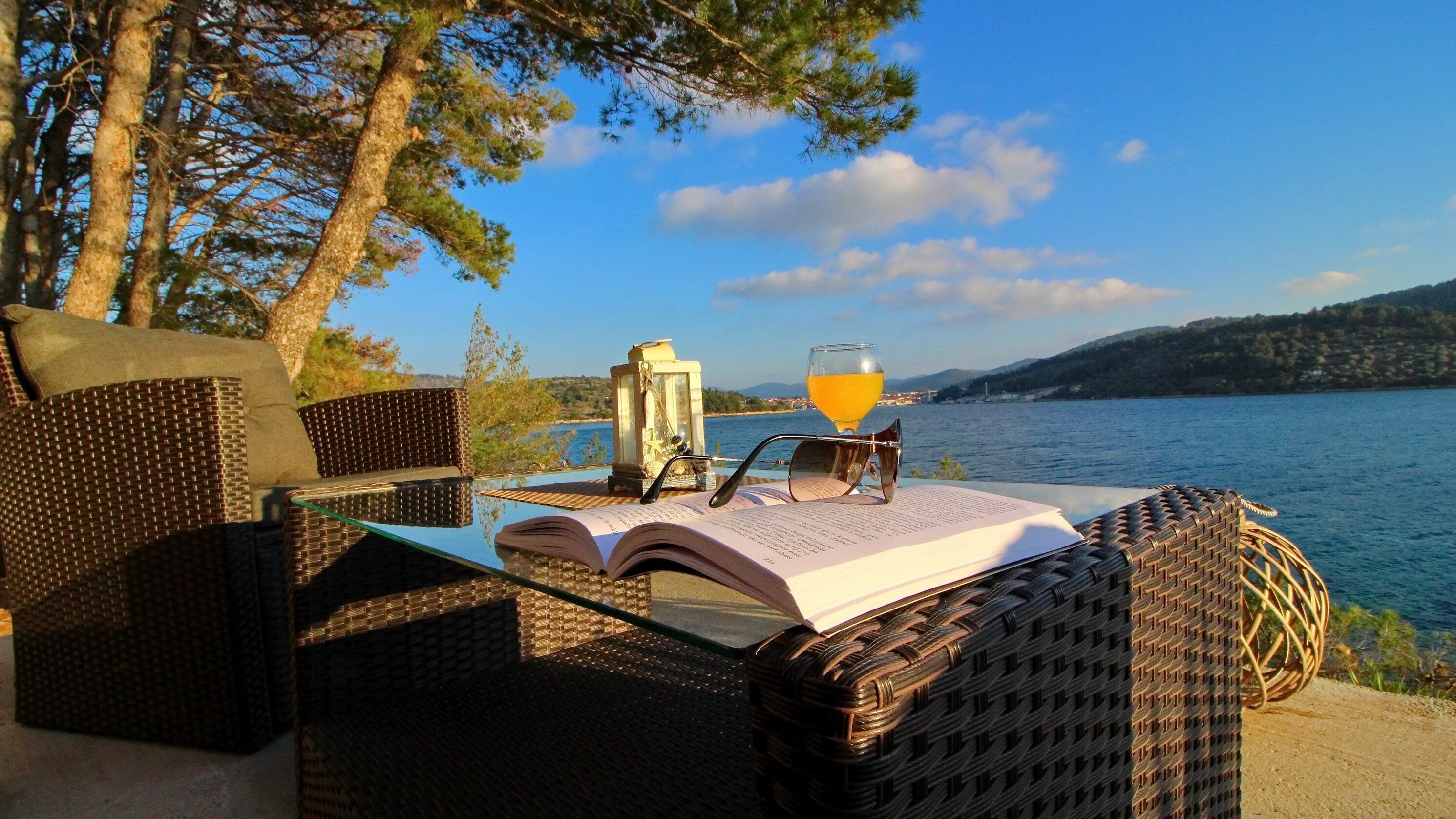 Wicker furniture, a book, a glass of orange juice, and a lantern by a lakeside.