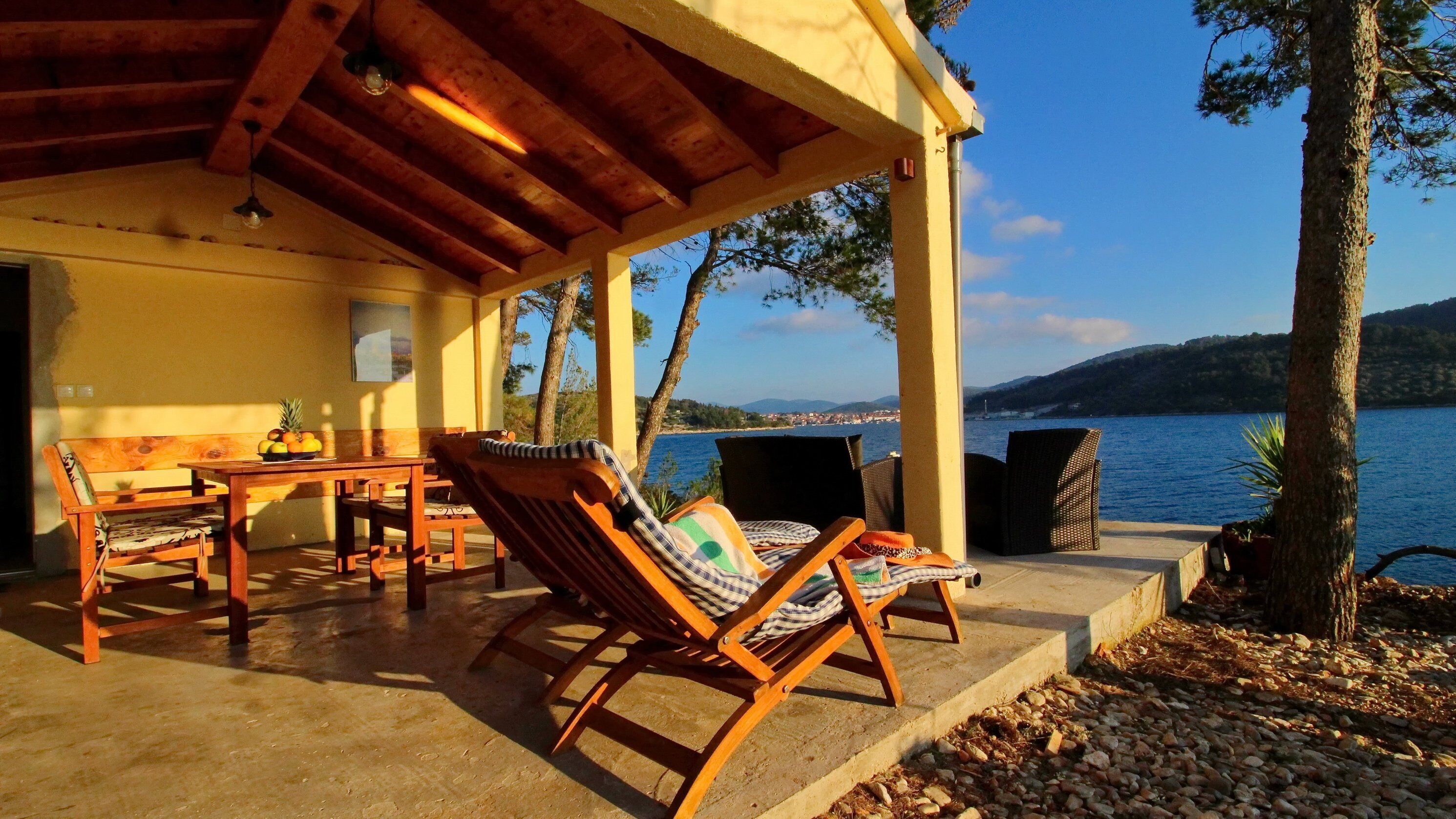 A wooden deck with a table and chairs, overlooking a body of water and hills.