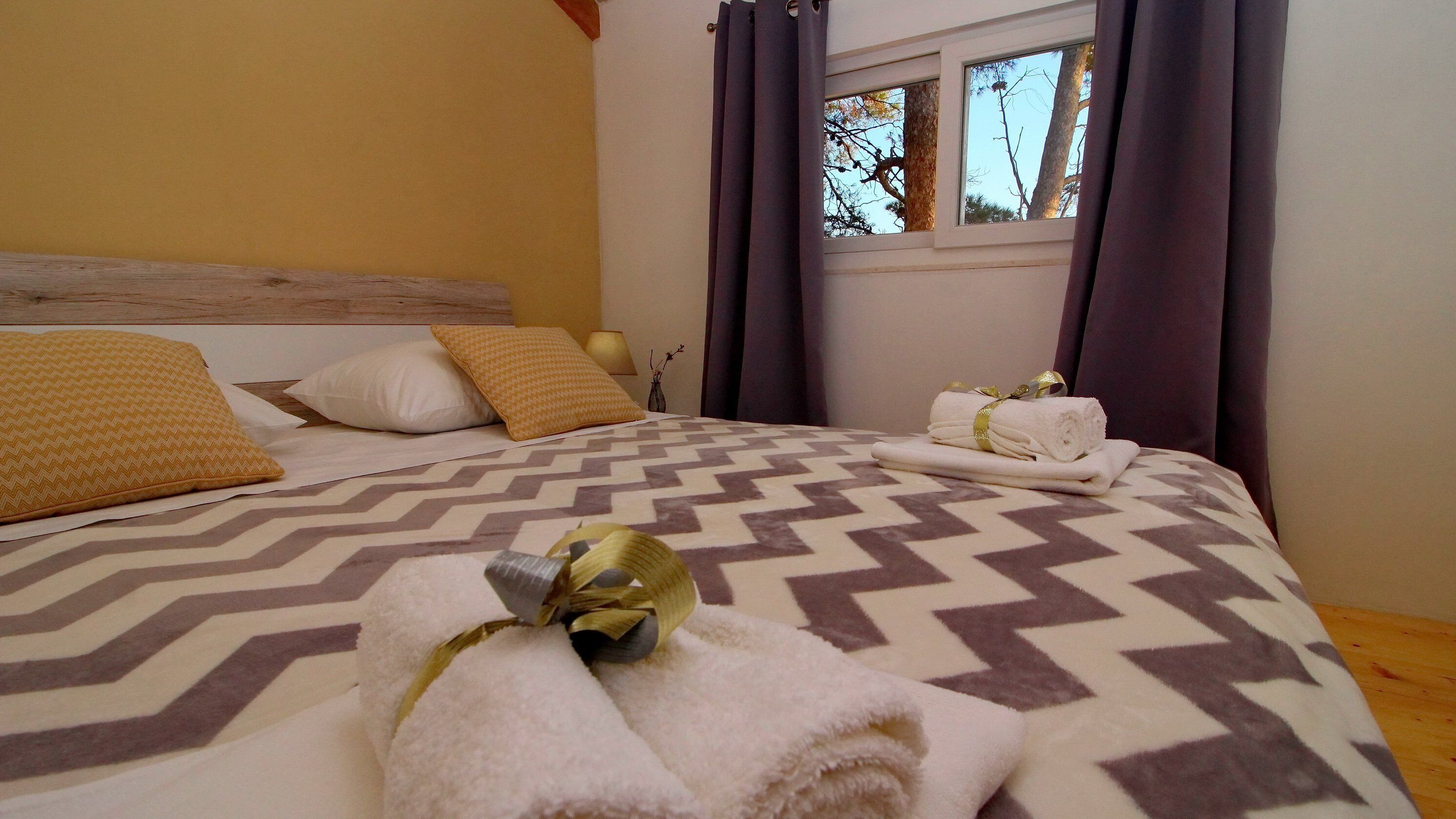 A neatly made bed with patterned bedding, a window with curtains, and a small table with towels and a vase.