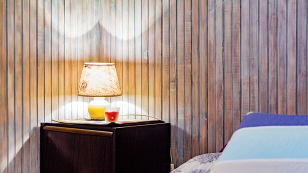 A wooden panel wall, a bedside table with a lamp, and a bed with a patterned bedspread. - Studio Apartment Silvana (Split)