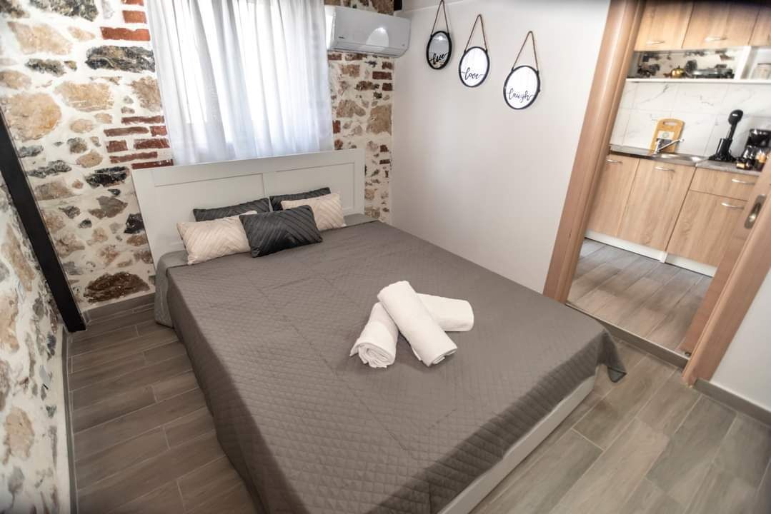 Une chambre avec un lit, une fenêtre avec des rideaux, un climatiseur fixé au mur et un coin cuisine visible à travers une porte ouverte.