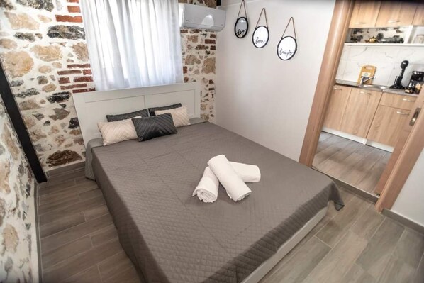 A bedroom with a bed, a window with curtains, a wall-mounted air conditioner, and a kitchen area visible through an open door.