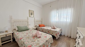 A bedroom with two beds, each with floral bedding and pillows, a nightstand, a bedside table, and a window with sheer curtains.