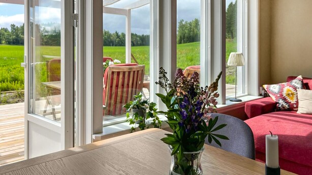 A room with large windows, a wooden table, a vase with flowers, a red sofa, and a lamp. - 2 bedroom amazing home in Säffle (Säffle)