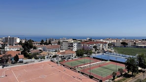 Uma quadra esportiva com campos de basquete e tênis, cercada por edifícios residenciais e uma vista desobstruída do mar.