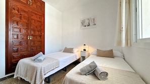 Quarto com duas camas, uma porta de madeira, uma obra de arte na parede e uma mesa de cabeceira com abajur.