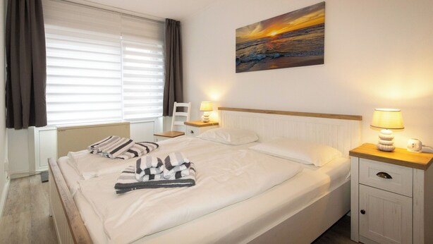 Een netjes opgemaakt bed met gestreepte en effen kussens, een nachtkastje met een lamp, en een schilderij van een zonsondergang aan de muur.