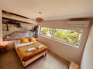 A bedroom with a large bed, a view of greenery through a window, a wall-mounted air conditioner, and a textured wall art piece.