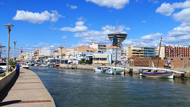 Stadsgezicht aan het water, met een opvallende toren, boten afgemeerd aan de steiger en diverse gebouwen op de achtergrond.
