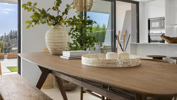 A modern kitchen with a wooden table, a large vase with greenery, and a view of the outdoors.