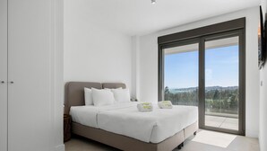 A bedroom with a bed, white bedding, a view through a sliding glass door, and a small bedside table.