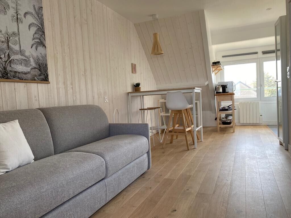 Ein modernes Wohnzimmer mit grauem Sofa, einem Essbereich mit weißem Tisch und Stühlen sowie einer Küchenzeile mit weißem Schrank und einer Pendelleuchte.