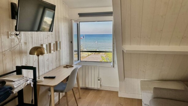 A room with a wooden interior, a desk with a chair, a TV mounted on the wall, and a view of the sea through a large window. - Studio Avec vue Mer, Centre de Langrune (Langrune-sur-Mer)