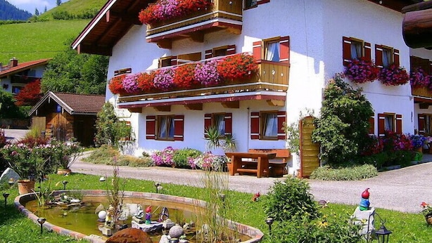 A multi-story building with balconies adorned with colorful flowers, a small pond, and a well-maintained garden. - Ferienwohnung Hochkalter, Dusche/wc, 57 qm, TV, Balkon, Obergeschoss (Ramsau bei Berchtesgaden)