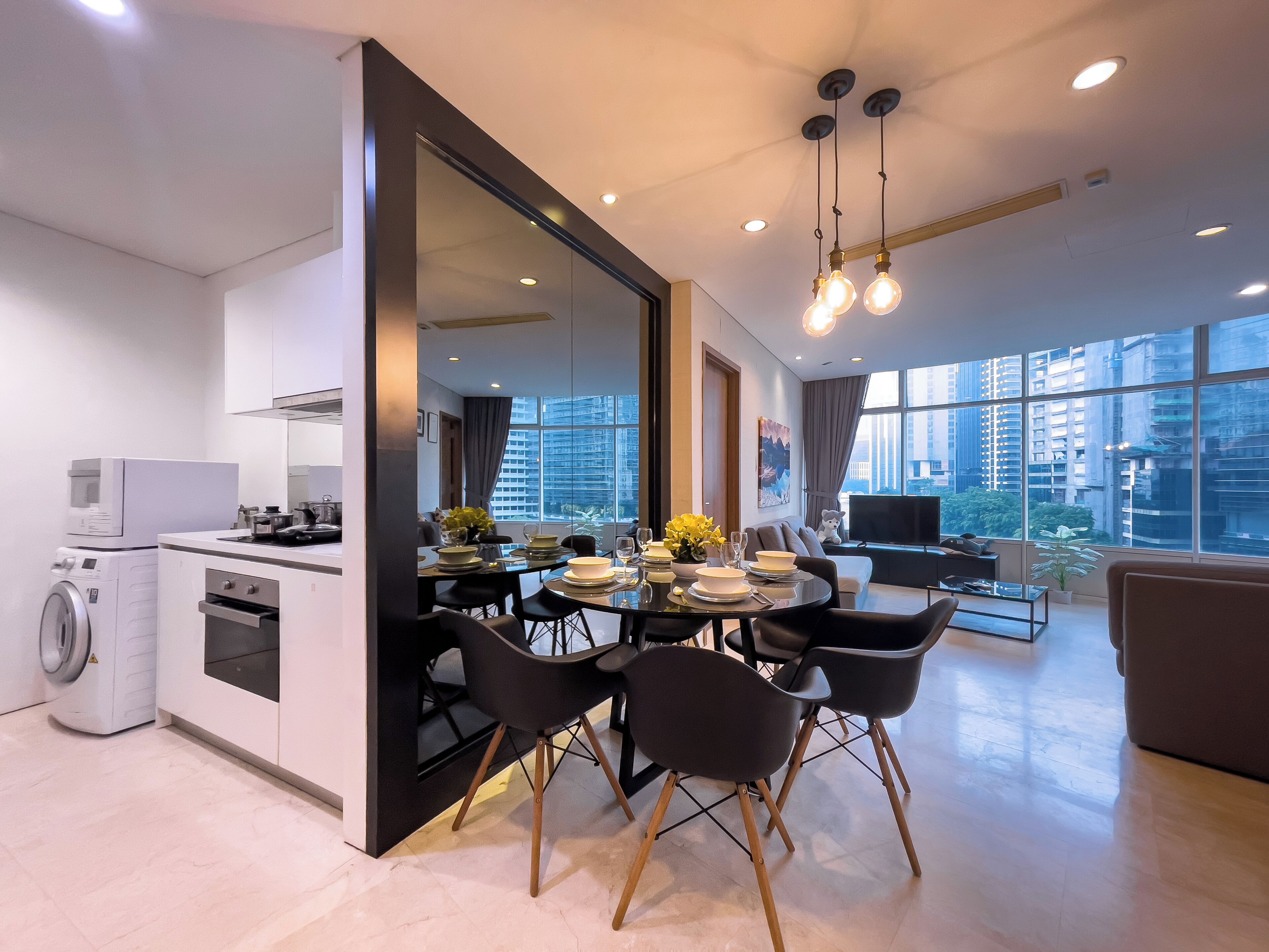 Una cocina moderna con horno y microondas integrados, un comedor con mesa redonda y sillas negras, y ventanas amplias con vistas a la ciudad.