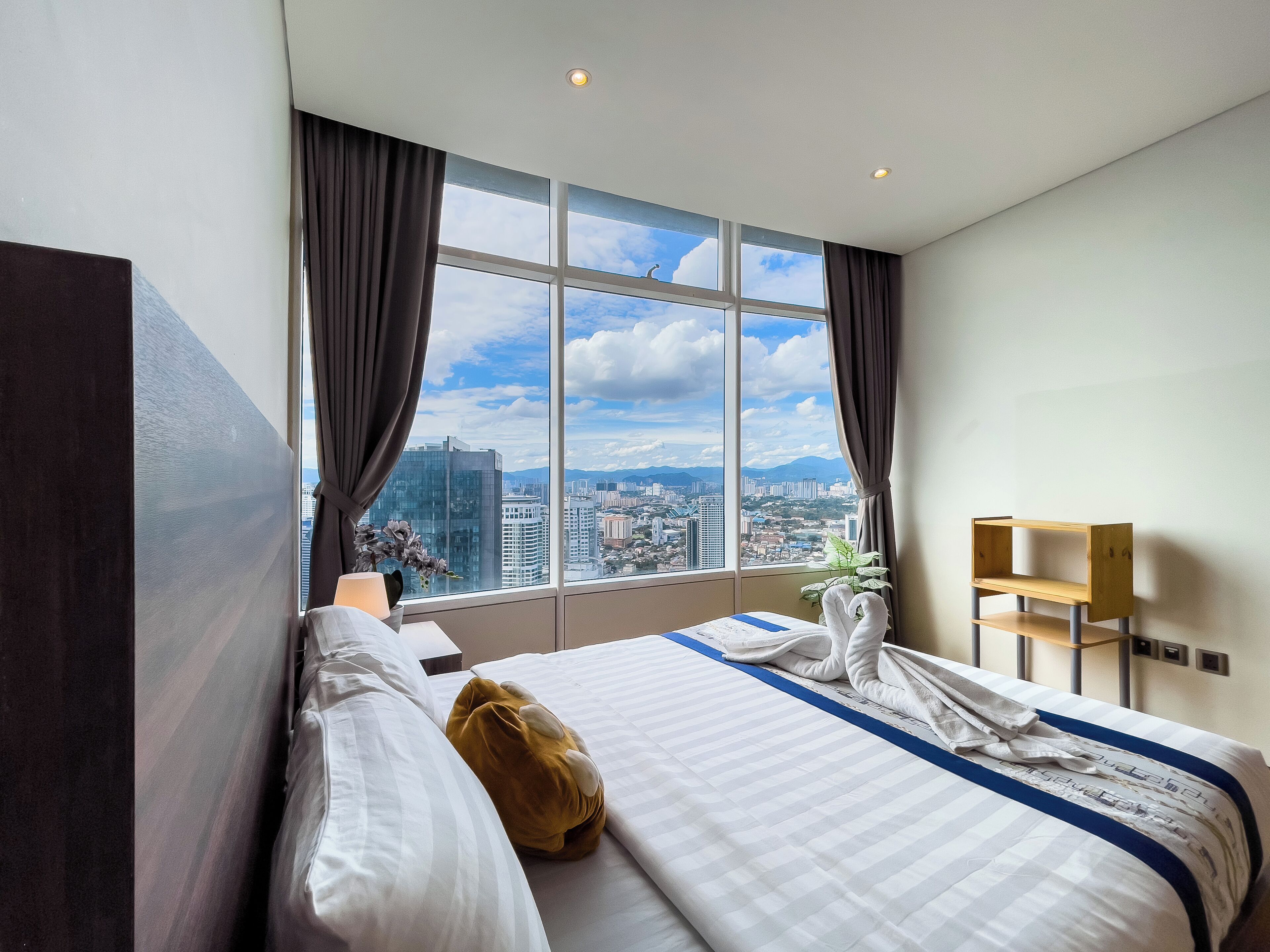 A hotel room with a large window, a bed, a wooden nightstand, and a view of the cityscape.