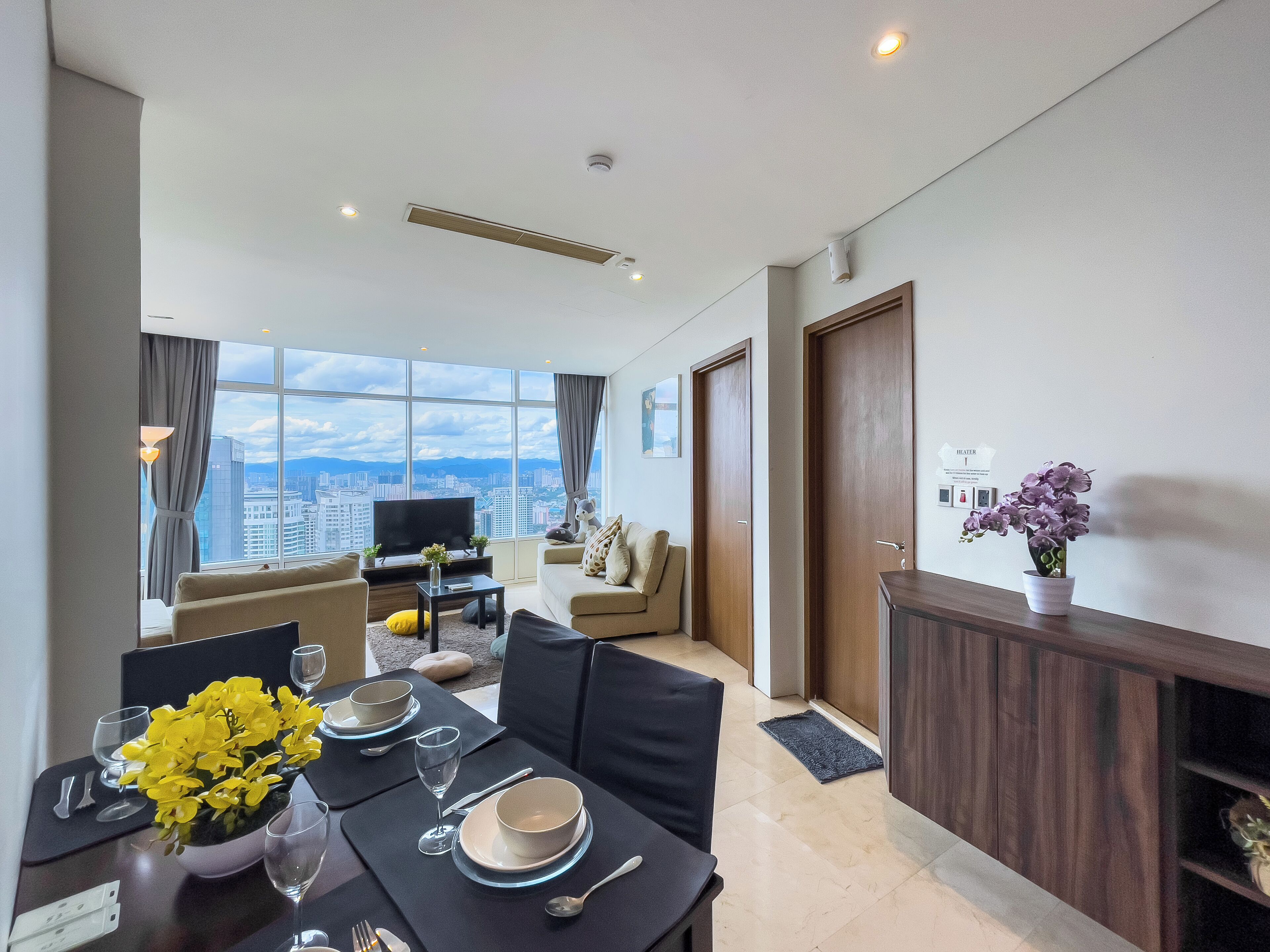A modern hotel room with a dining area, a sofa, a TV, and a view of the cityscape.