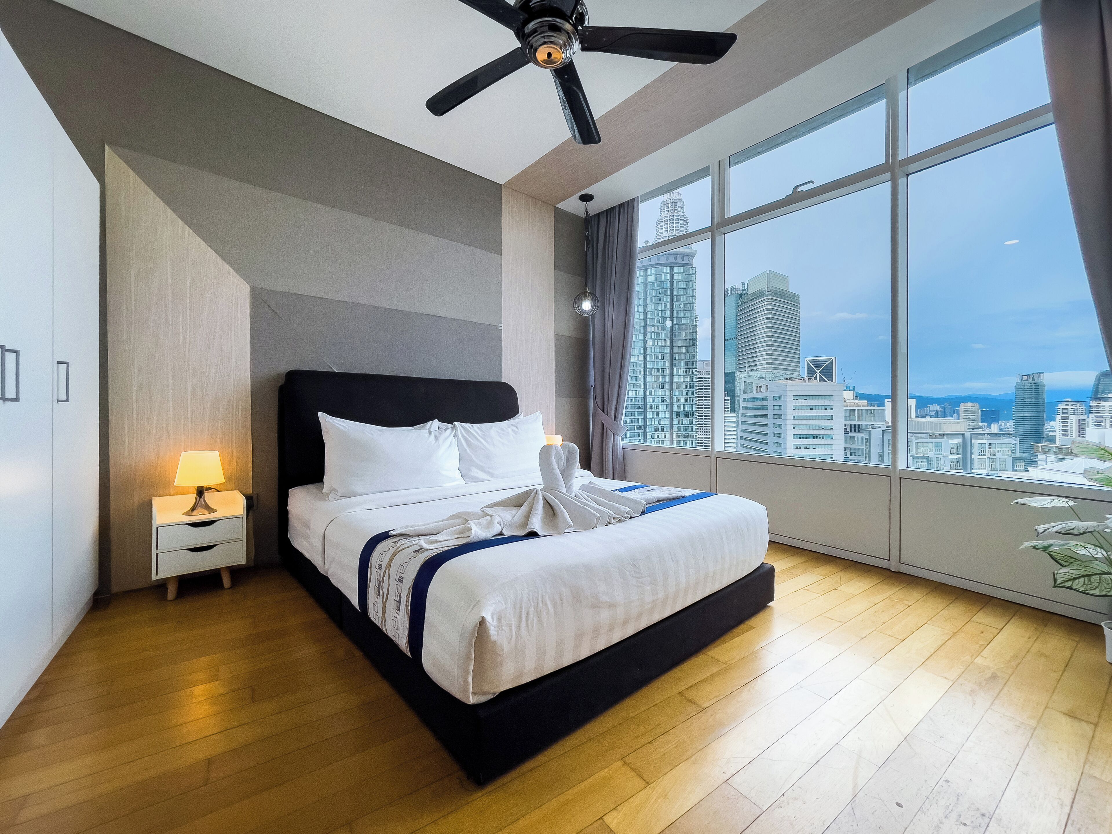 A modern hotel room with a large bed, a ceiling fan, a nightstand with a lamp, and a view of the city skyline through large windows.