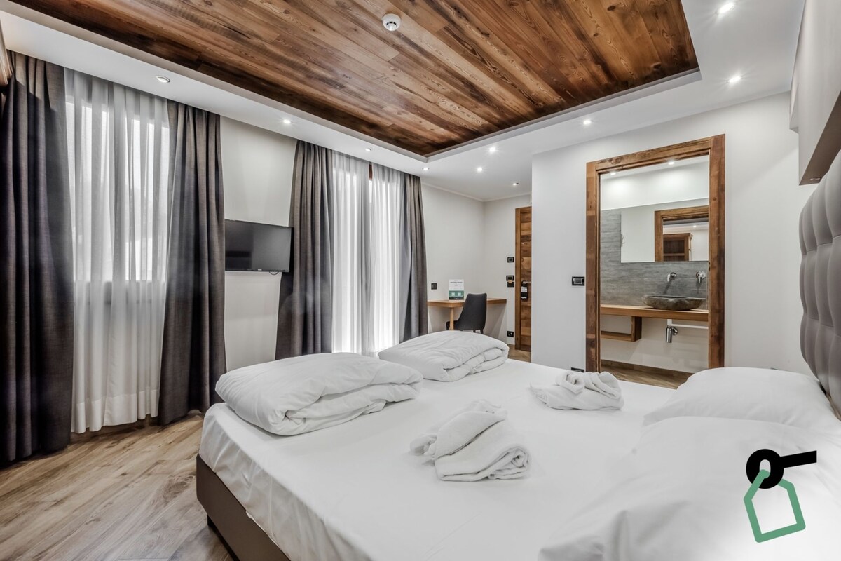 A modern hotel room with a wooden ceiling, a large bed with white linens, a bathroom with a mirror and sink, and a desk with a chair.