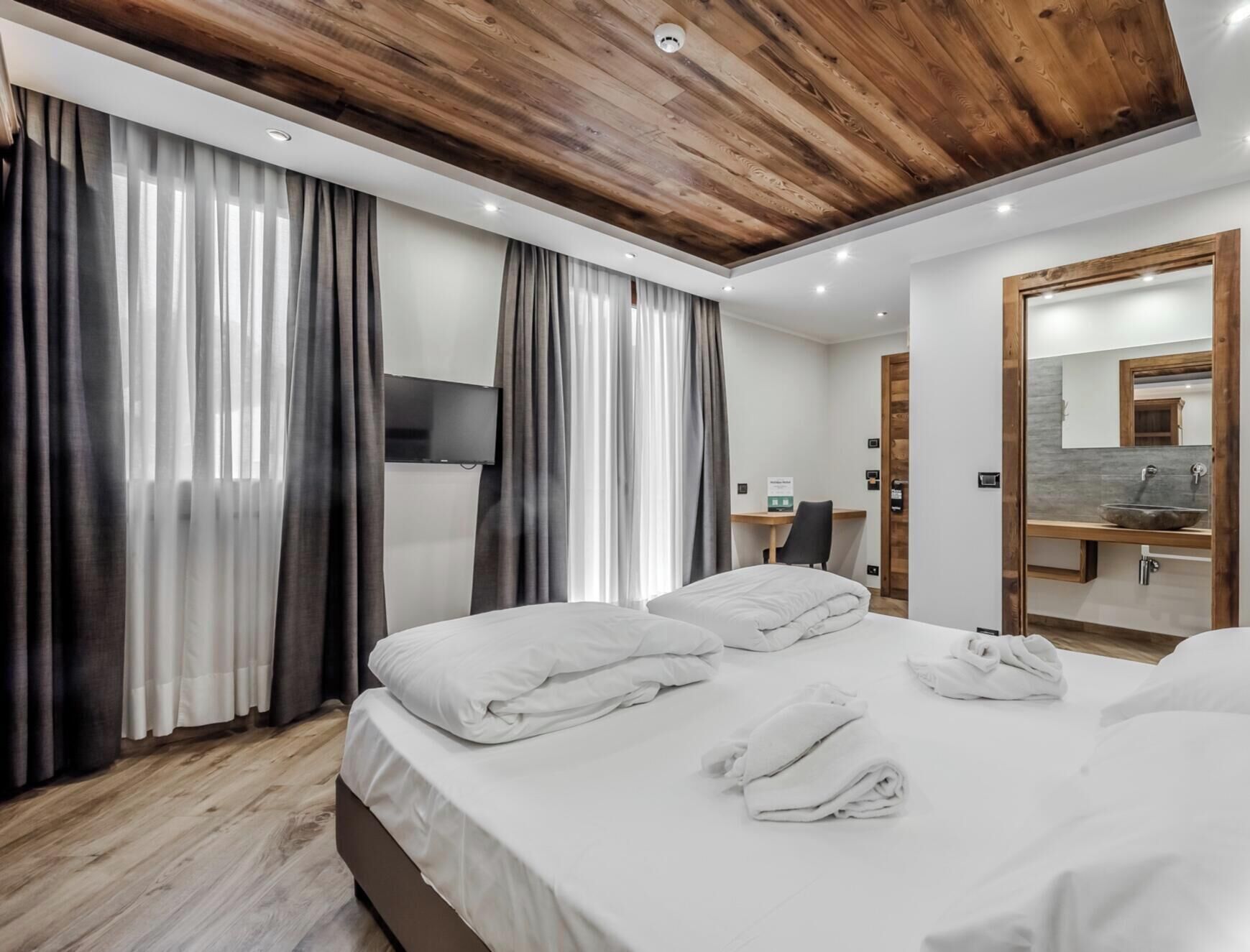 Une chambre d’hôtel moderne avec un plafond en bois, un grand lit, un bureau et une salle de bain équipée d’un miroir et d’un lavabo.