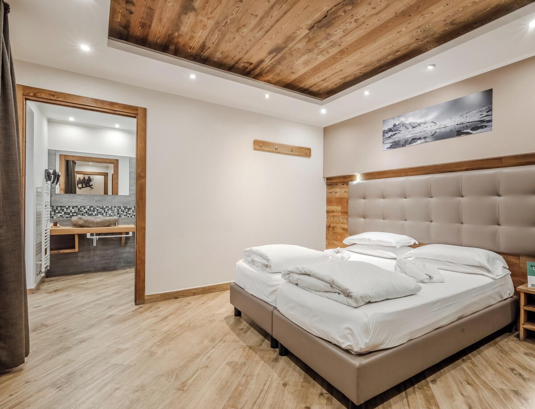 Un dormitorio con techo de madera, una cama con ropa de cama blanca, cabecera de madera y un baño con espejo y lavamanos.