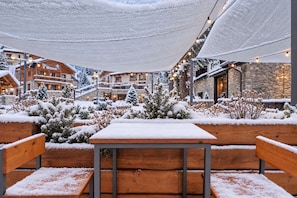 Un área de estar al aire libre cubierta de nieve, con bancos y mesas de madera, rodeada de plantas y árboles cargados de nieve, bajo una estructura de techo.