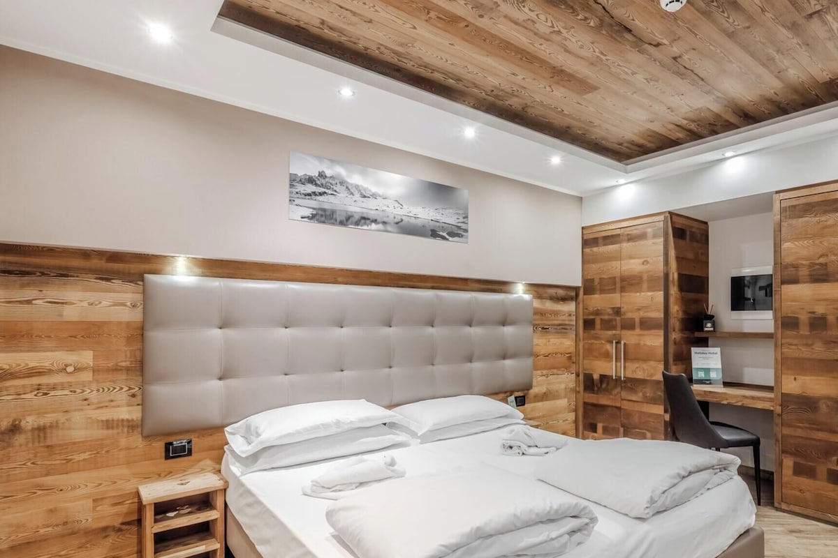 A modern bedroom with a wooden headboard, a bed with white linens, a small bedside table, and a wooden wardrobe.
